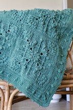 A teal green crocheted throw blanket with a textured design draped over a light brown wooden chair in a home. setting.