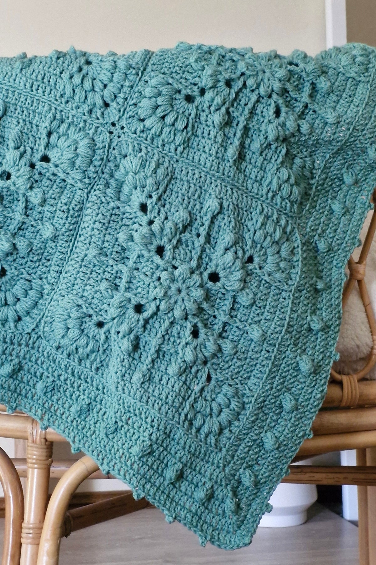 A teal green crocheted throw blanket with a textured design draped over a light brown wooden chair in a home. setting.