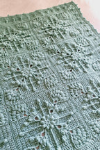 A close-up of a handmade crocheted blanket, featuring intricate floral and snowflake-like lace patterns in a muted sage green color.