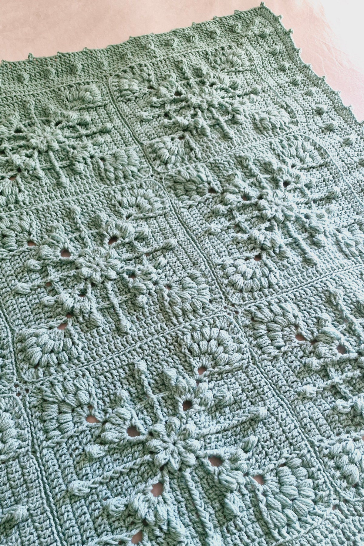 A close-up of a handmade crocheted blanket, featuring intricate floral and snowflake-like lace patterns in a muted sage green color.