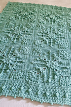A handmade crocheted blanket in a light mint green color, featuring intricate snowflake and floral granny square patterns, laid flat against a white fabric background.