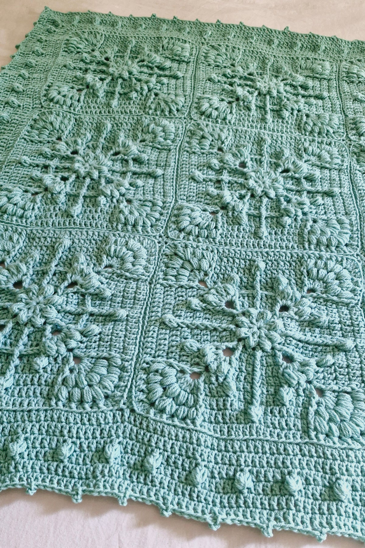 A handmade crocheted blanket in a light mint green color, featuring intricate snowflake and floral granny square patterns, laid flat against a white fabric background.