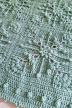 A close-up of a handmade crocheted blanket in a soft sage green color, featuring intricate floral and snowflake-like patterns.