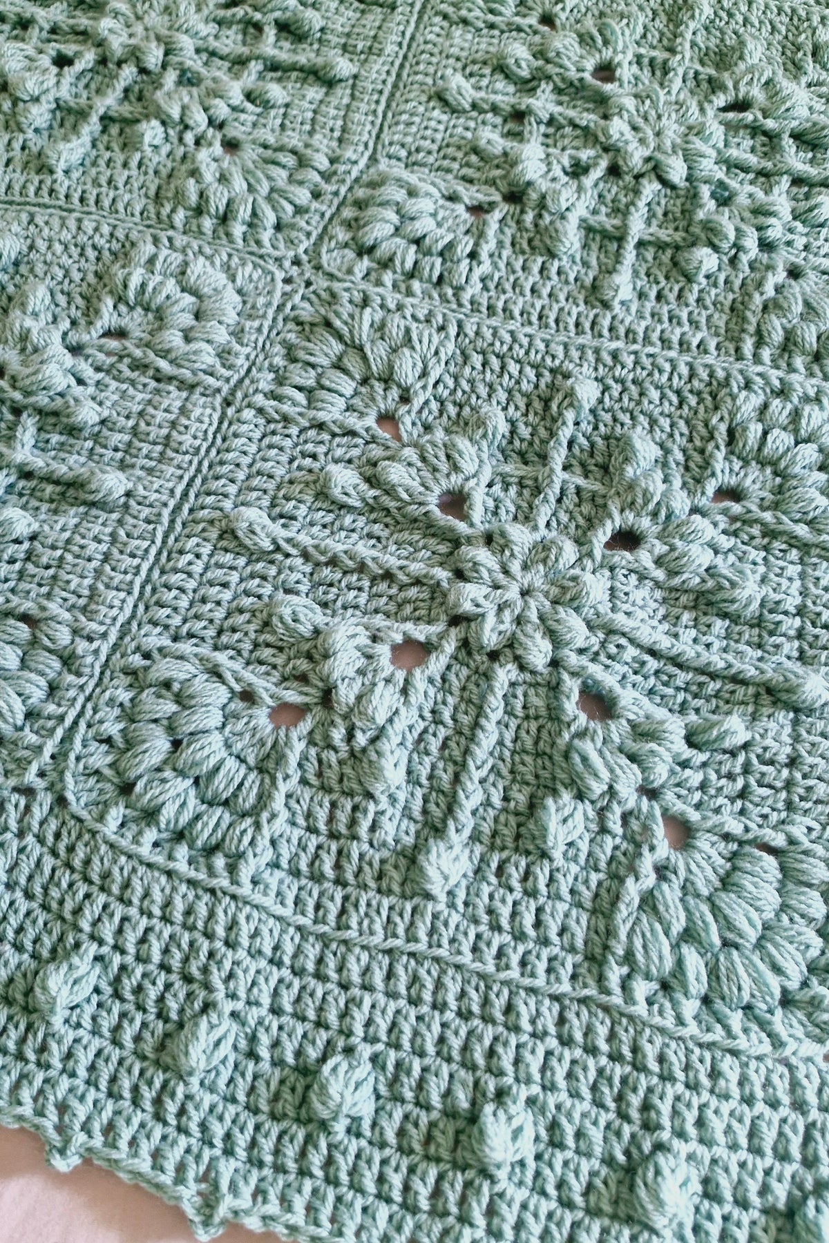 A close-up of a handmade crocheted blanket in a soft sage green color, featuring intricate floral and snowflake-like patterns.