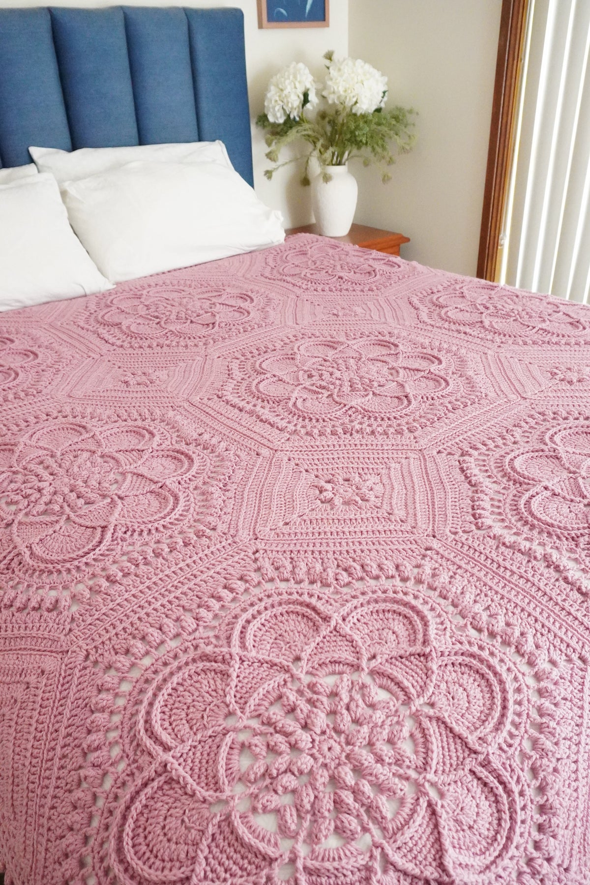 A pink crocheted blanket with intricate floral and geometric patterns draped over a bed in a bedroom setting.