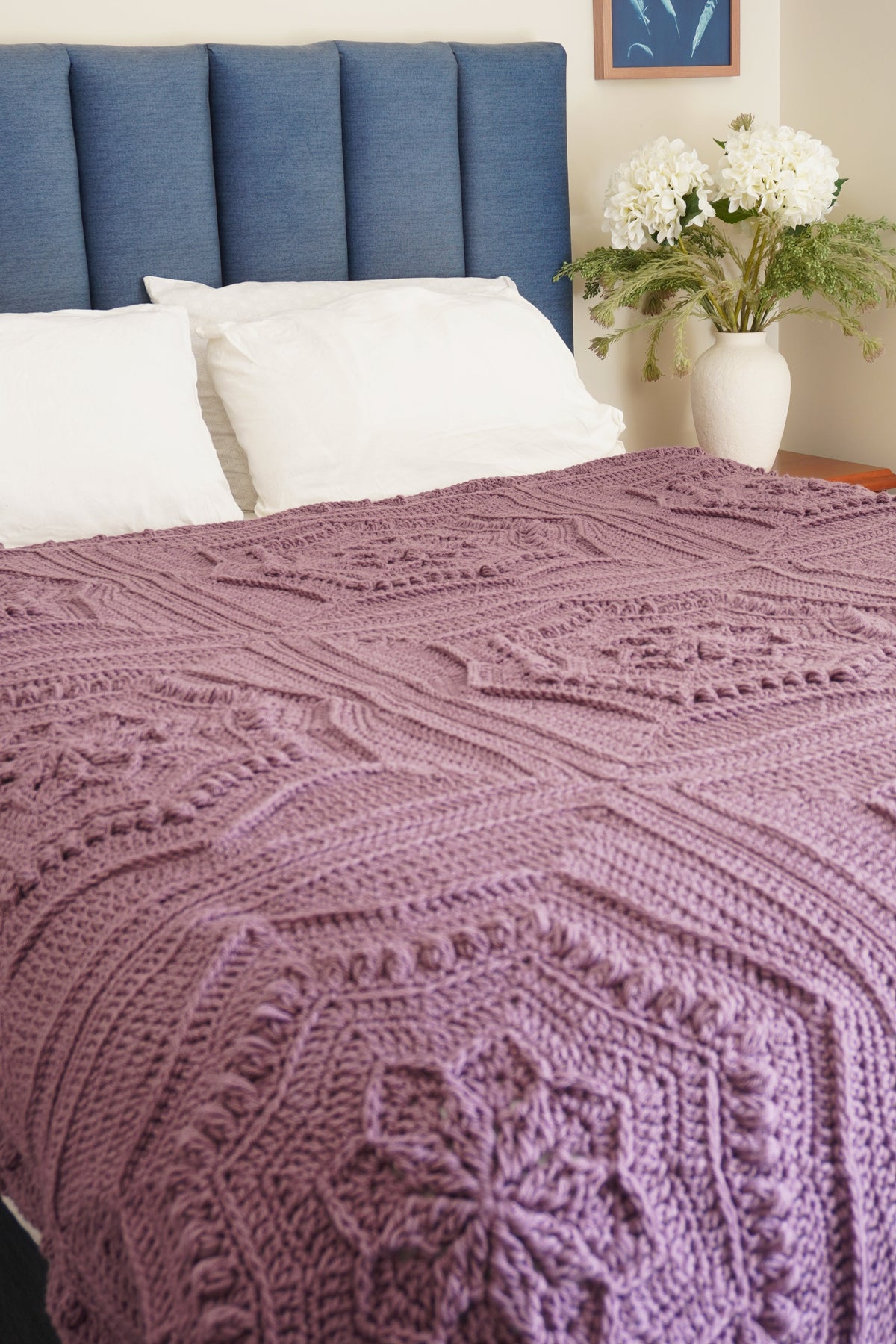 A textured, hand-crocheted blanket in a dusty mauve-purple color is spread across a bed with white pillows and a blue upholstered headboard in a bedroom setting.