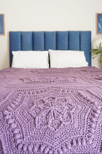 A close-up of a purple crocheted blanket with a star mandala pattern styled on a bed with a blue upholstered headboard and white pillows.