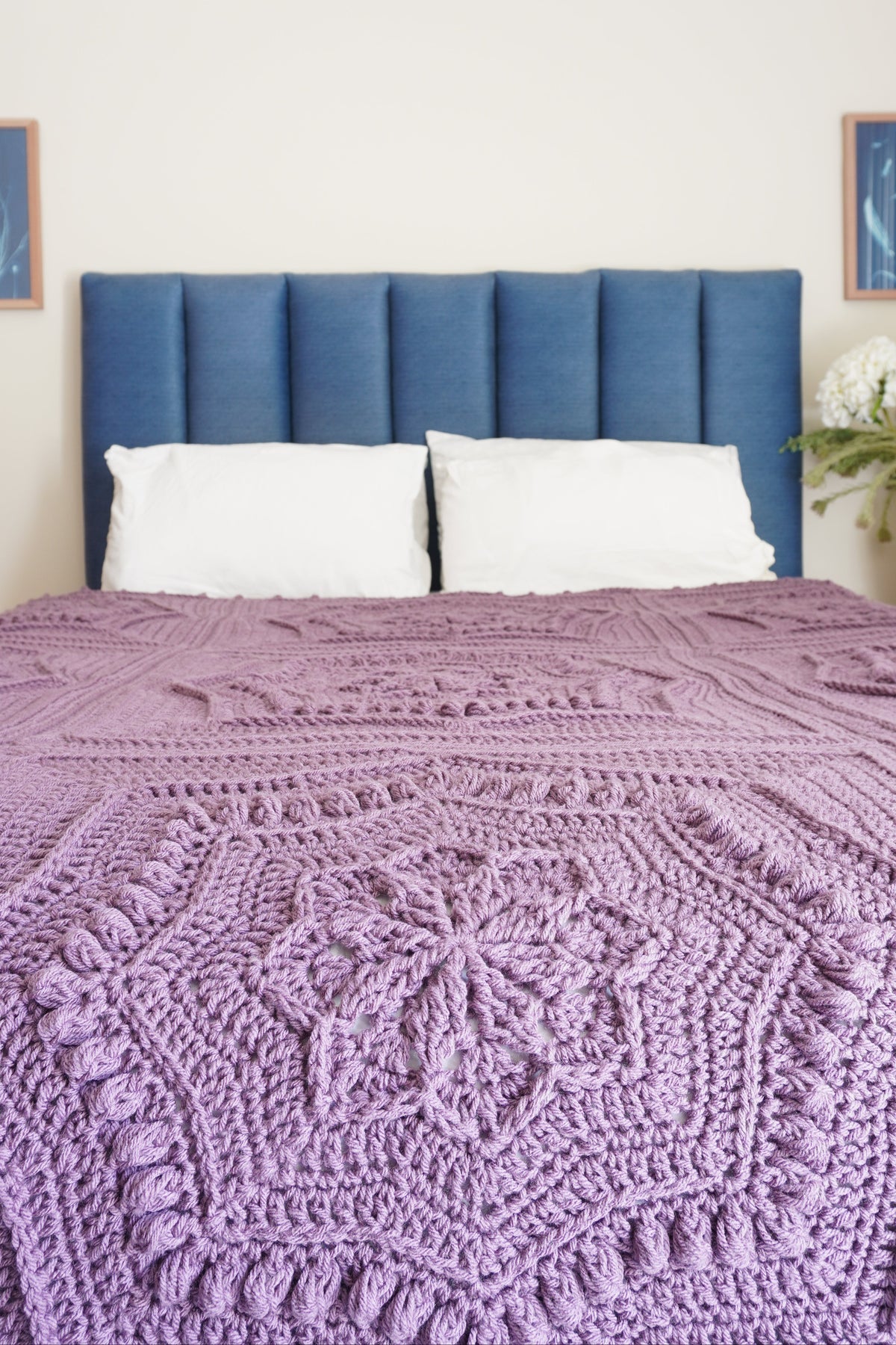 A close-up of a purple crocheted blanket with a star mandala pattern styled on a bed with a blue upholstered headboard and white pillows.