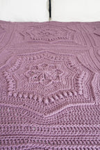 A close-up of a handmade crocheted blanket in a dusty mauve-purple color, featuring intricate geometric and floral lace patterns.