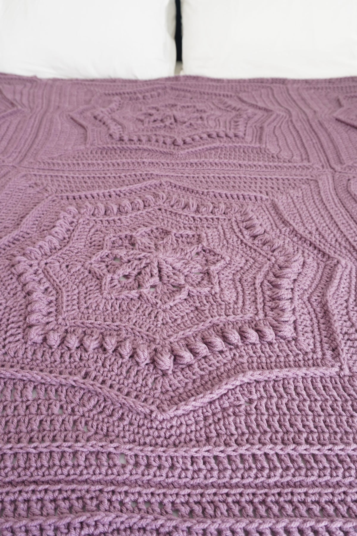 A close-up of a handmade crocheted blanket in a dusty mauve-purple color, featuring intricate geometric and floral lace patterns.