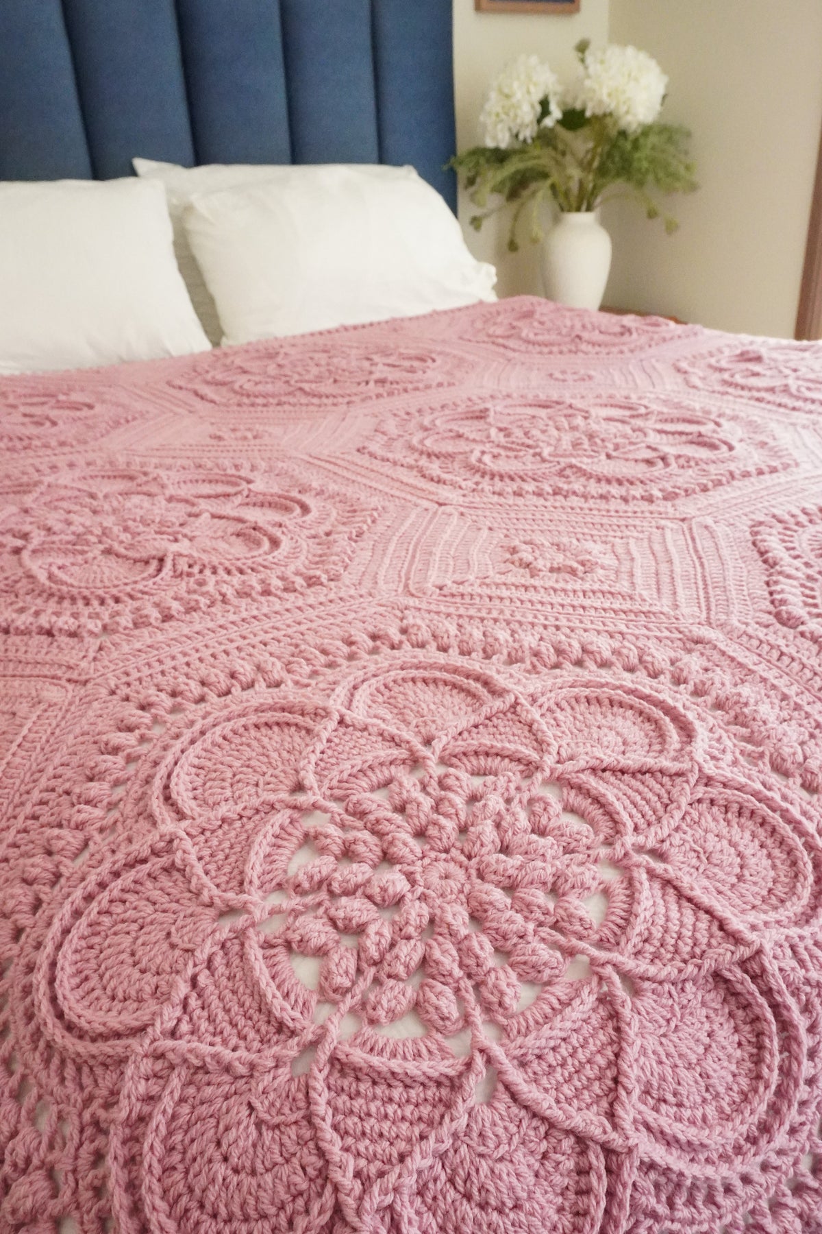 A pink crocheted blanket with intricate floral and geometric patterns draped over a bed in a bedroom setting, with a blue upholstered headboard, white pillows, and a vase of flowers in the background.