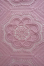 A close-up of a pink crocheted blanket with an intricate floral and geometric lace pattern, filling the entire frame.