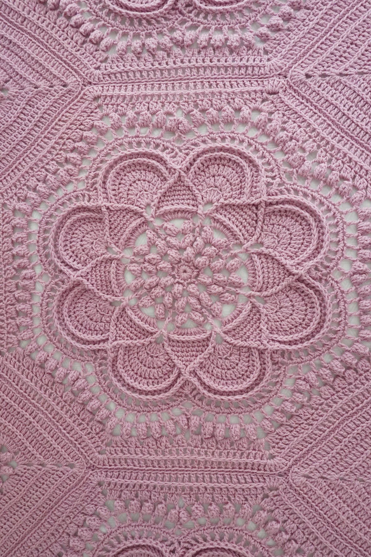 A close-up of a pink crocheted blanket with an intricate floral and geometric lace pattern, filling the entire frame.