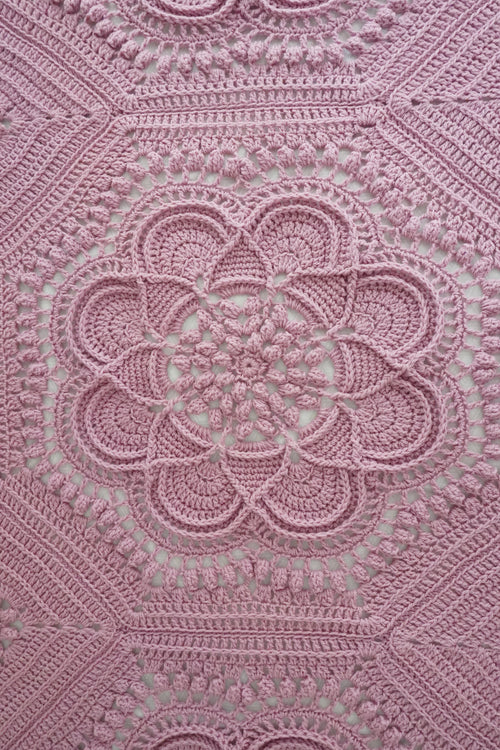 A close-up of a pink crocheted blanket with an intricate floral and geometric lace pattern, filling the entire frame.
