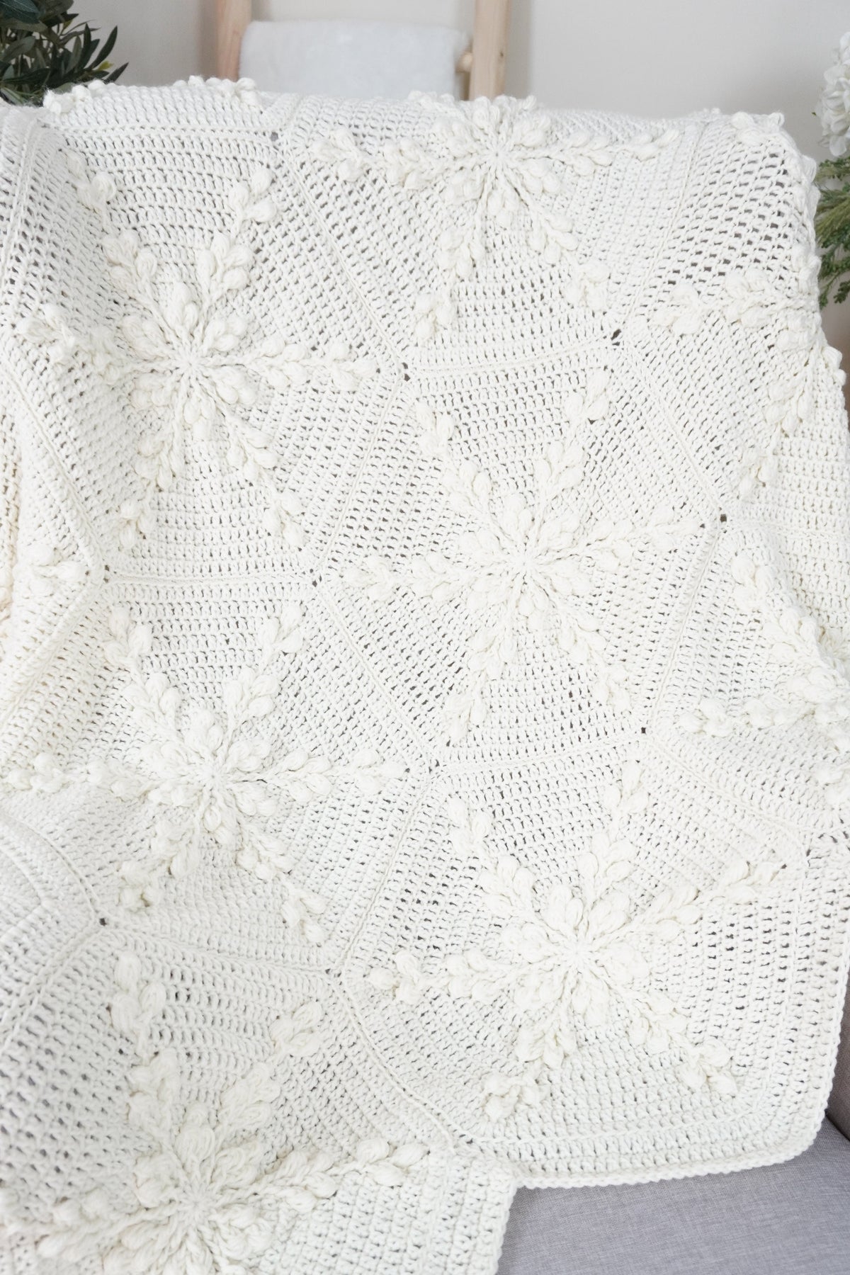An off-white crocheted blanket with a snowflake design draped over a light gray sofa in a home setting.