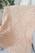 A beige crocheted throw blanket with intricate circular mandala patterns draped over a light gray sofa in a bright, modern room.