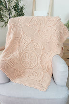 A beige-colored crocheted throw blanket with intricate circular and floral patterns draped over a light gray upholstered armchair in a bright, modern room.