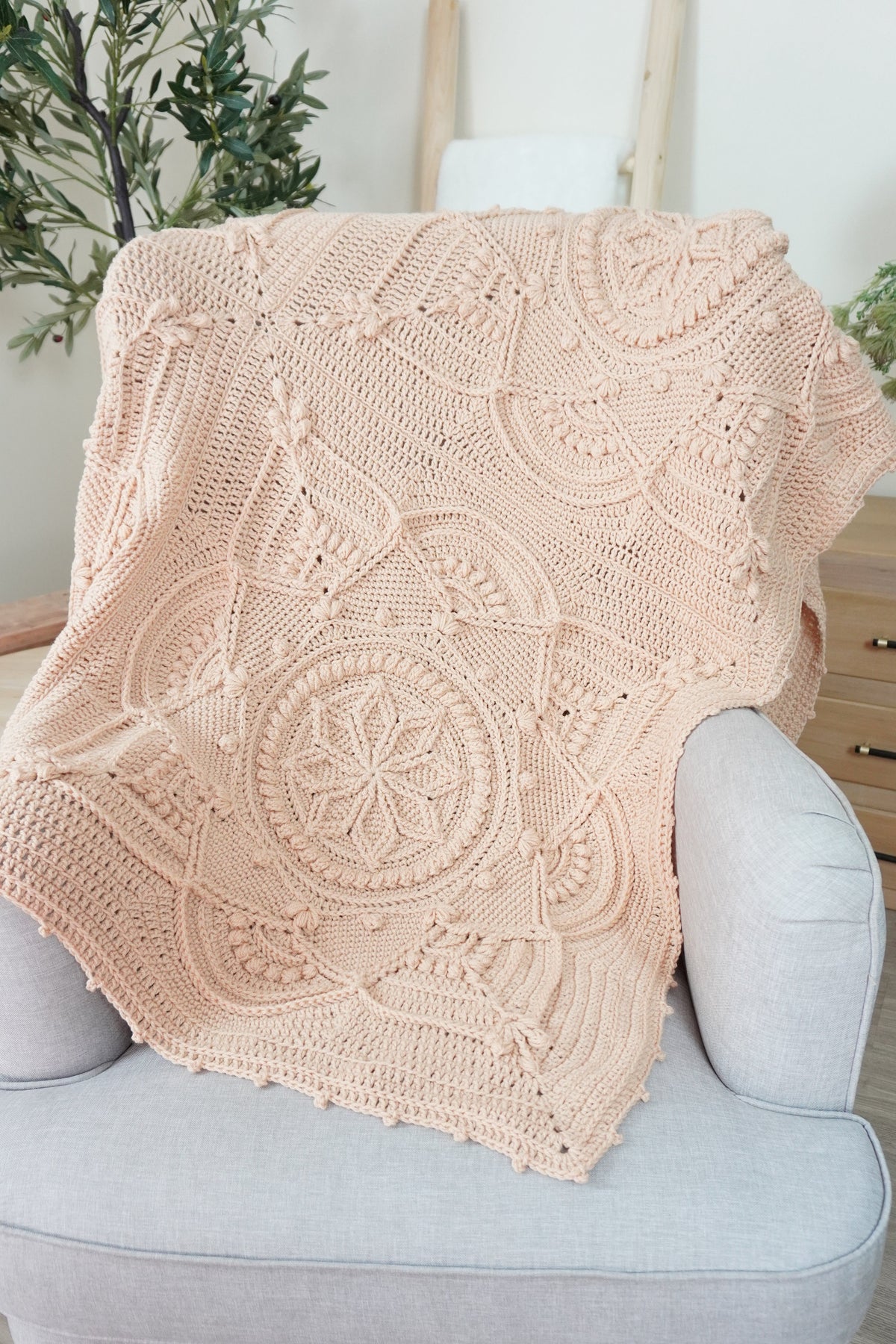 A beige-colored crocheted throw blanket with intricate circular and floral patterns draped over a light gray upholstered armchair in a bright, modern room.