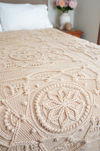 A close-up of a beige hand-crocheted blanket draped over a bed in a softly lit bedroom.