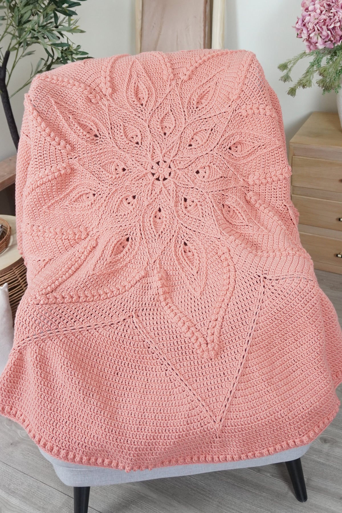 A peach-pink crocheted throw blanket with an intricate leafy mandala pattern draped over a light gray armchair in a cozy room setting.