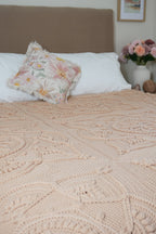 A close-up of a textured, beige-colored crochet blanket with a floral pattern draped over a bed, styled with white pillows and a matching floral accent pillow in a bedroom setting.