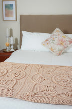 A beige-colored crocheted blanket with an intricate lace-like pattern is draped over the foot of a bed with white bedding, a beige headboard, and a floral accent pillow.