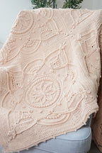 A light beige crocheted throw blanket with textured pattern draped over a light gray upholstered armchair, with a green plant in the background.