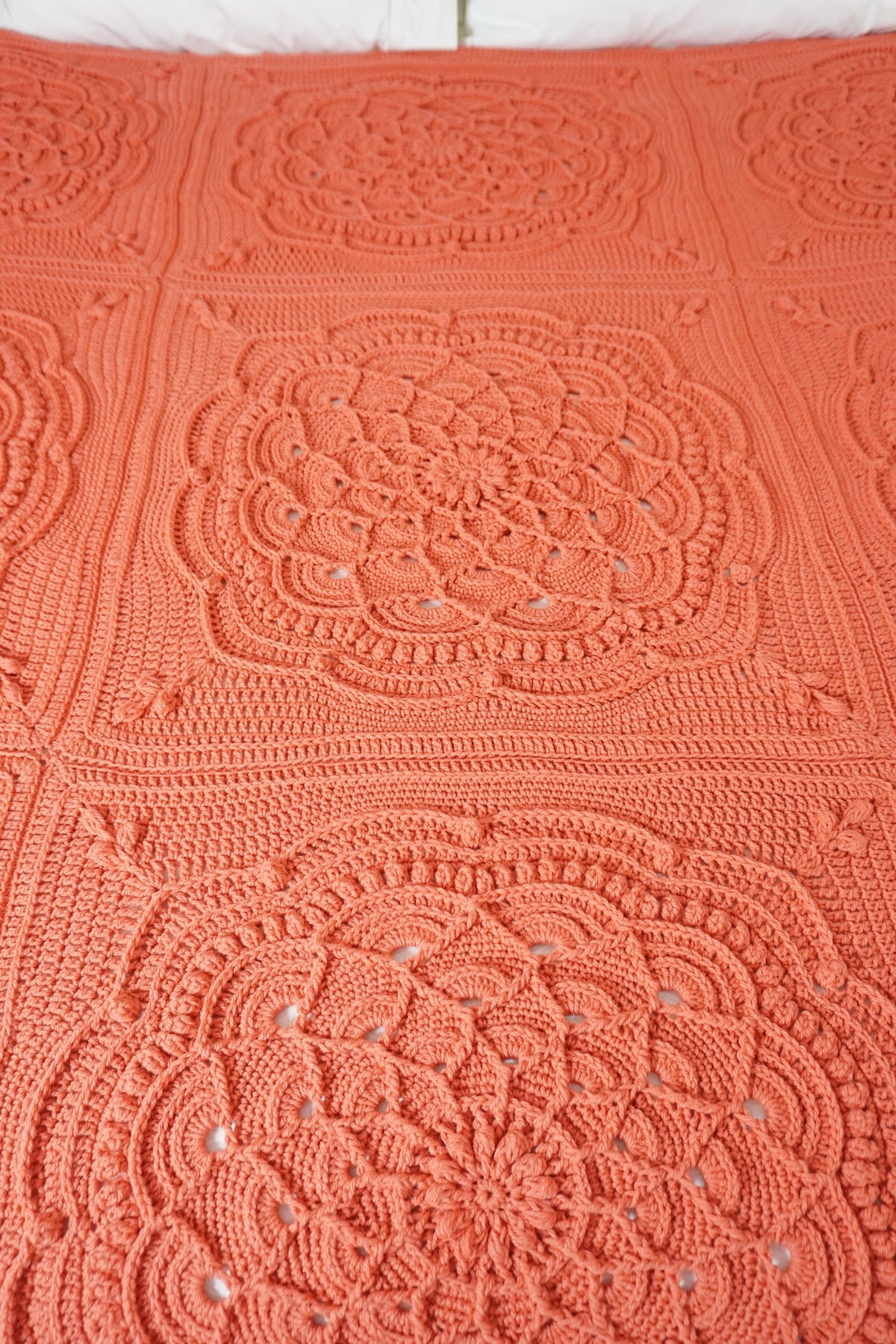A close-up of a crocheted blanket featuring large square panels with intricate circular lace-like medallion patterns in a warm coral-orange color.