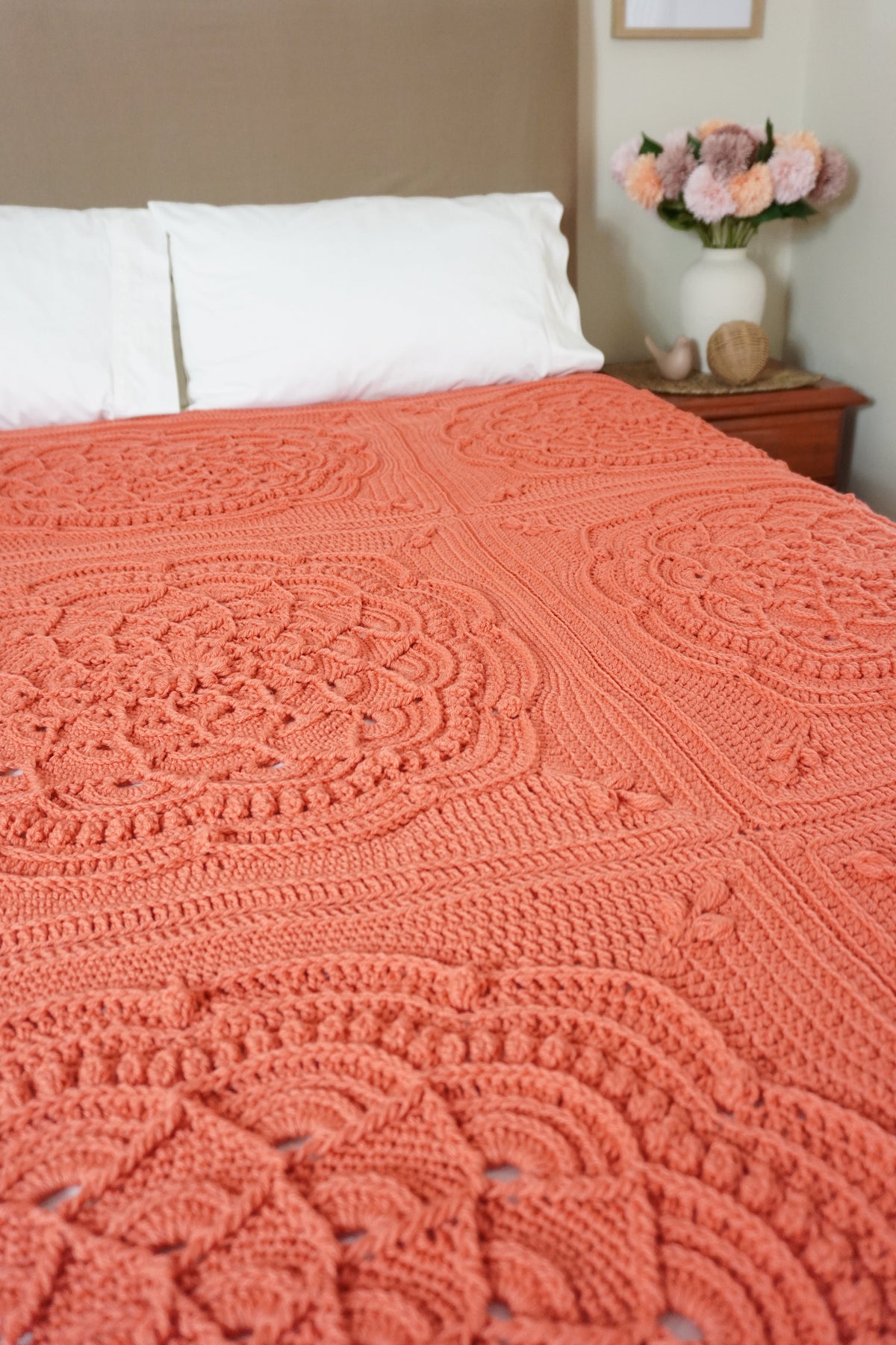 A handmade coral orange crochet blanket with intricate circular mandala patterns is spread across a bed in a bedroom setting, featuring a beige headboard, white pillows, and a bedside table with flowers.
