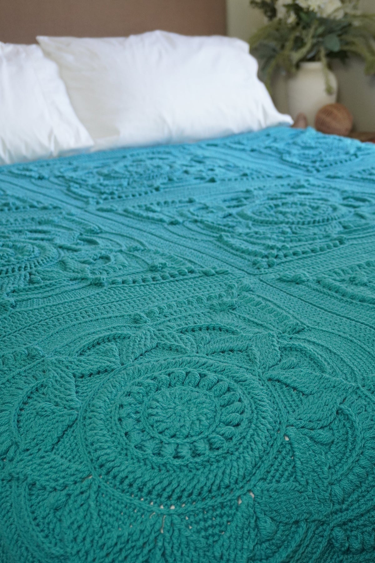 A teal crocheted blanket with intricate circular and floral lace patterns draped over a bed with white pillows in a bedroom setting.