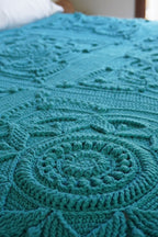 A close-up of a teal crocheted blanket with intricate circular and geometric patterns draped over a bed.