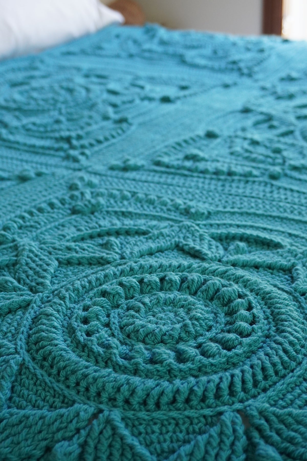A close-up of a teal crocheted blanket with intricate circular and geometric patterns draped over a bed.