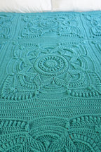 A close-up of a teal crocheted blanket with intricate floral and geometric lace patterns, laid flat on a bed with white pillows visible at the top.