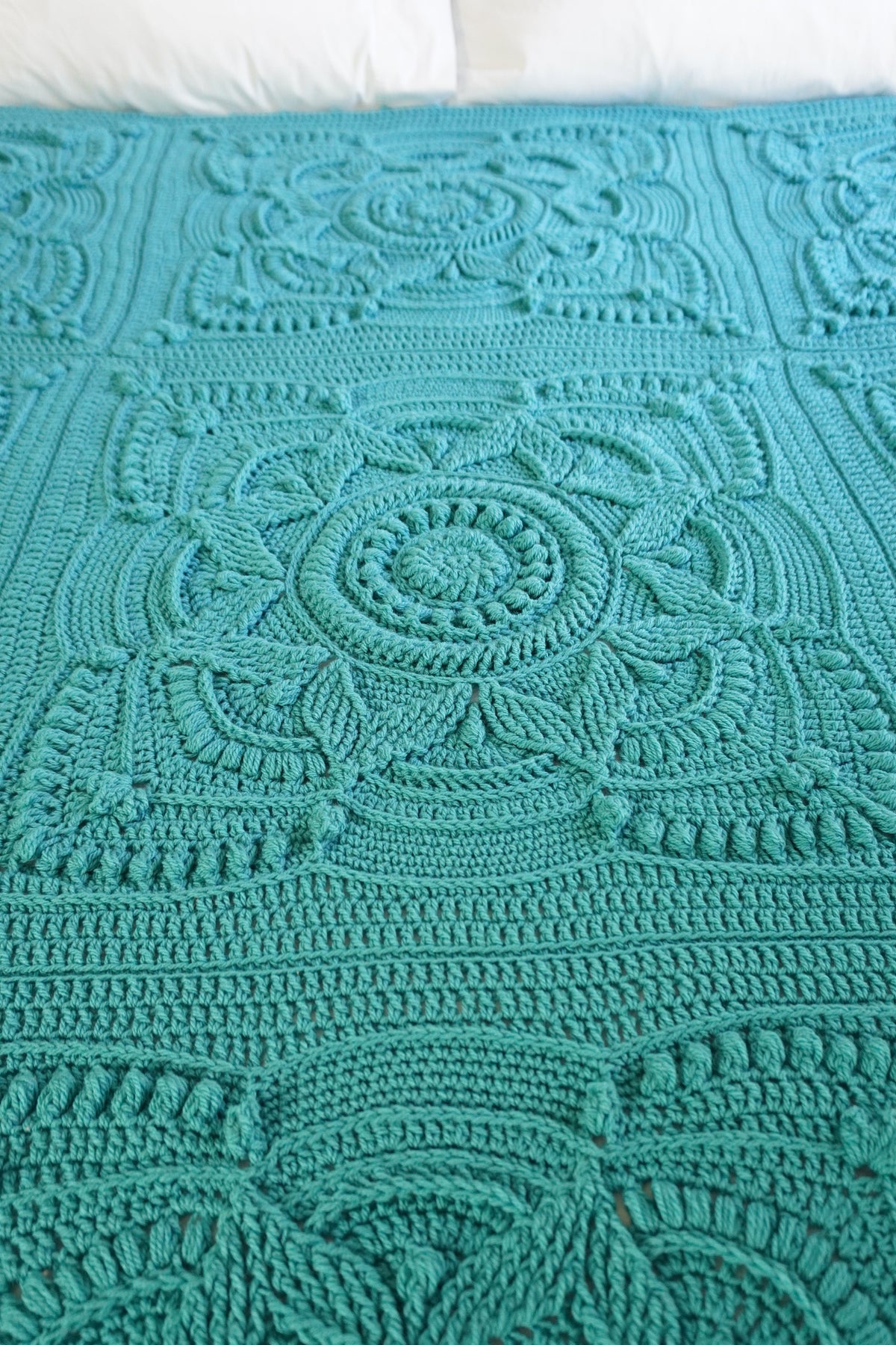 A close-up of a teal crocheted blanket with intricate floral and geometric lace patterns, laid flat on a bed with white pillows visible at the top.