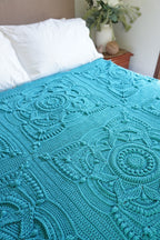 A turquoise blue crocheted blanket with intricate floral and geometric patterns draped over a bed with white pillows in a bedroom setting.
