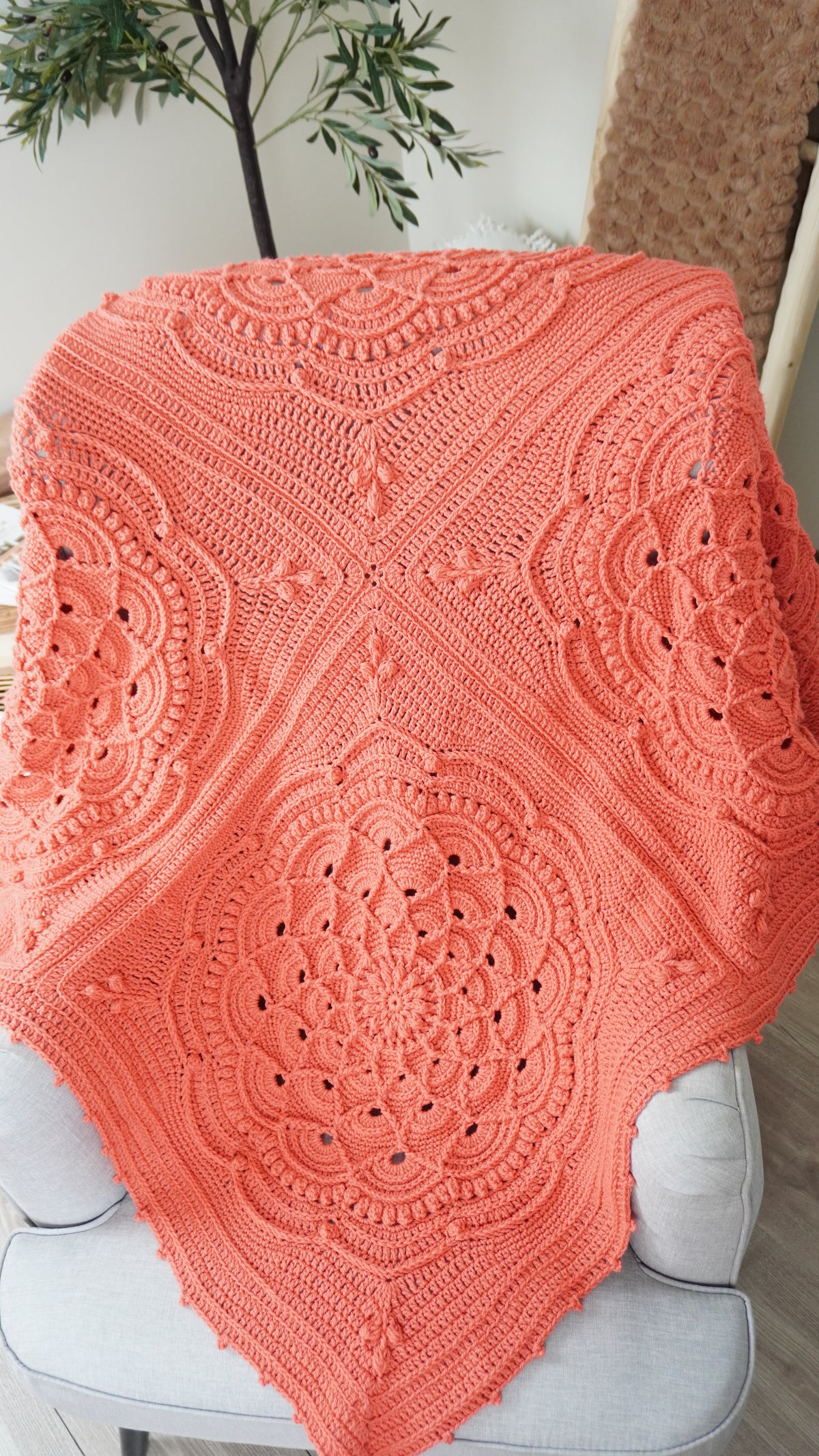 A large coral orange crochet shawl with intricate mandala and floral patterns draped over a light gray upholstered chair in a home setting.