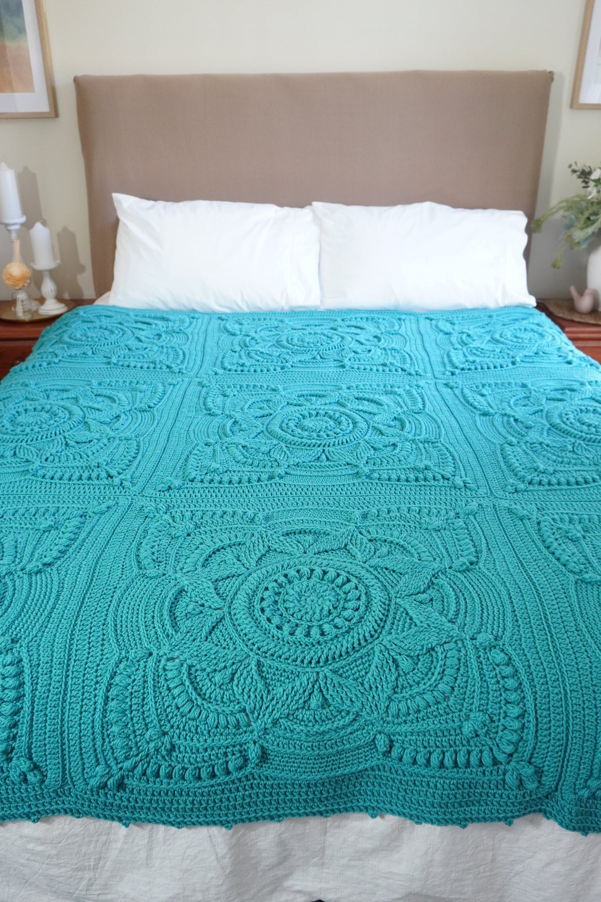 A bright turquoise crocheted blanket with intricate mandala and floral patterns draped over a bed with white pillows and a beige headboard.