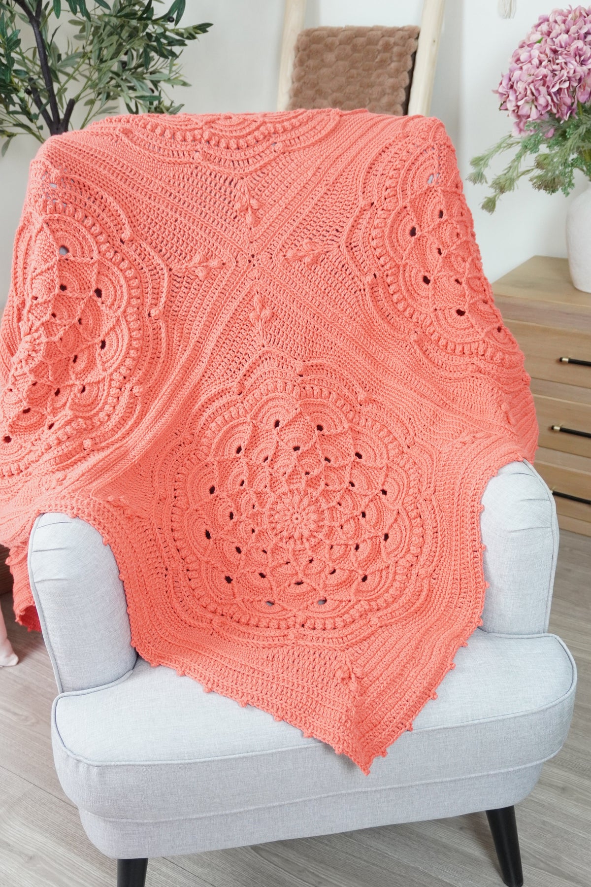 A coral crocheted blanket with intricate lace and mandala patterns draped over a light gray upholstered armchair in a bright, modern room.