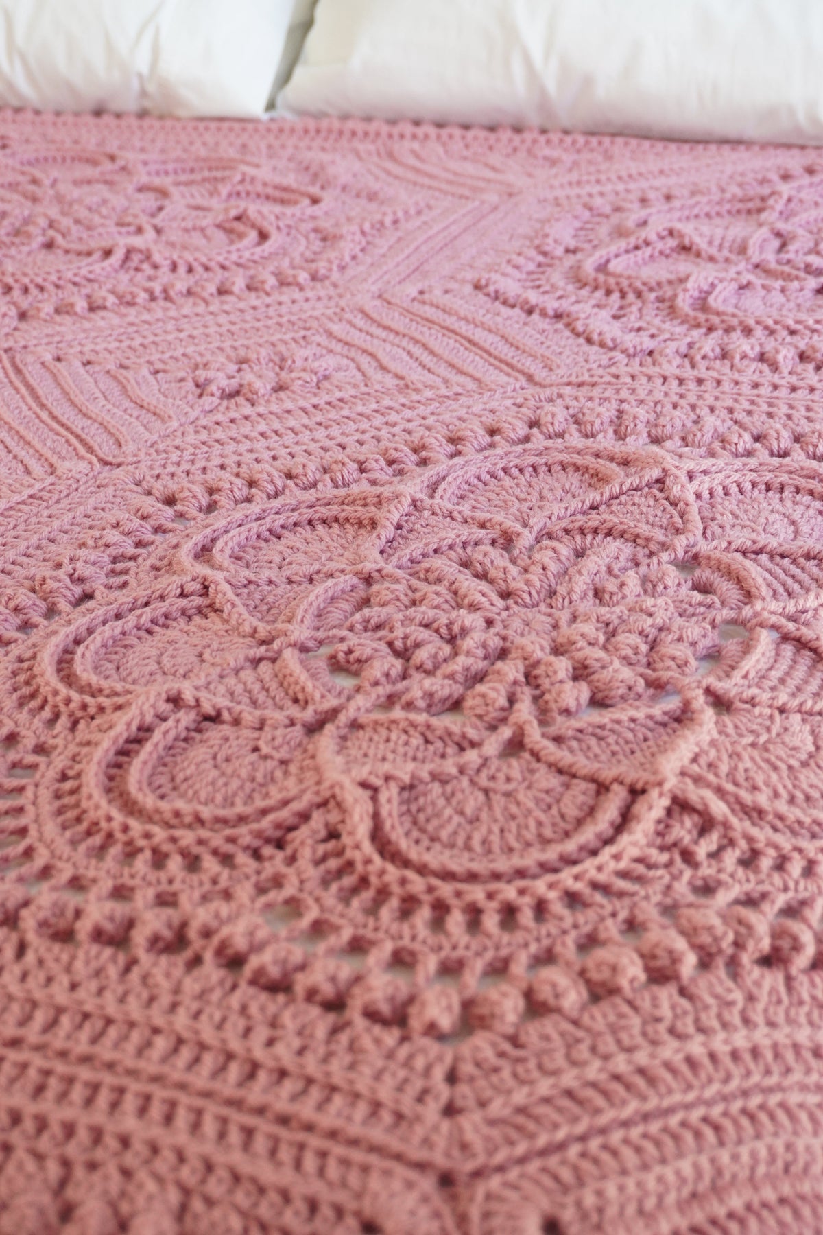 A close-up of a dusty pink crocheted blanket with intricate floral and geometric lace patterns, styled on a bed with white pillows visible at the top.