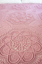 A close-up of a pink crocheted blanket with intricate floral and geometric lace patterns, laid out on a bed with white pillows visible at the top.