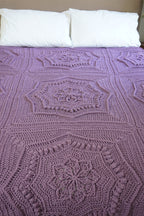 A handmade crocheted blanket with a textured octagonal pattern in dusty purple, styled on a bed with two white pillows against a beige wall.