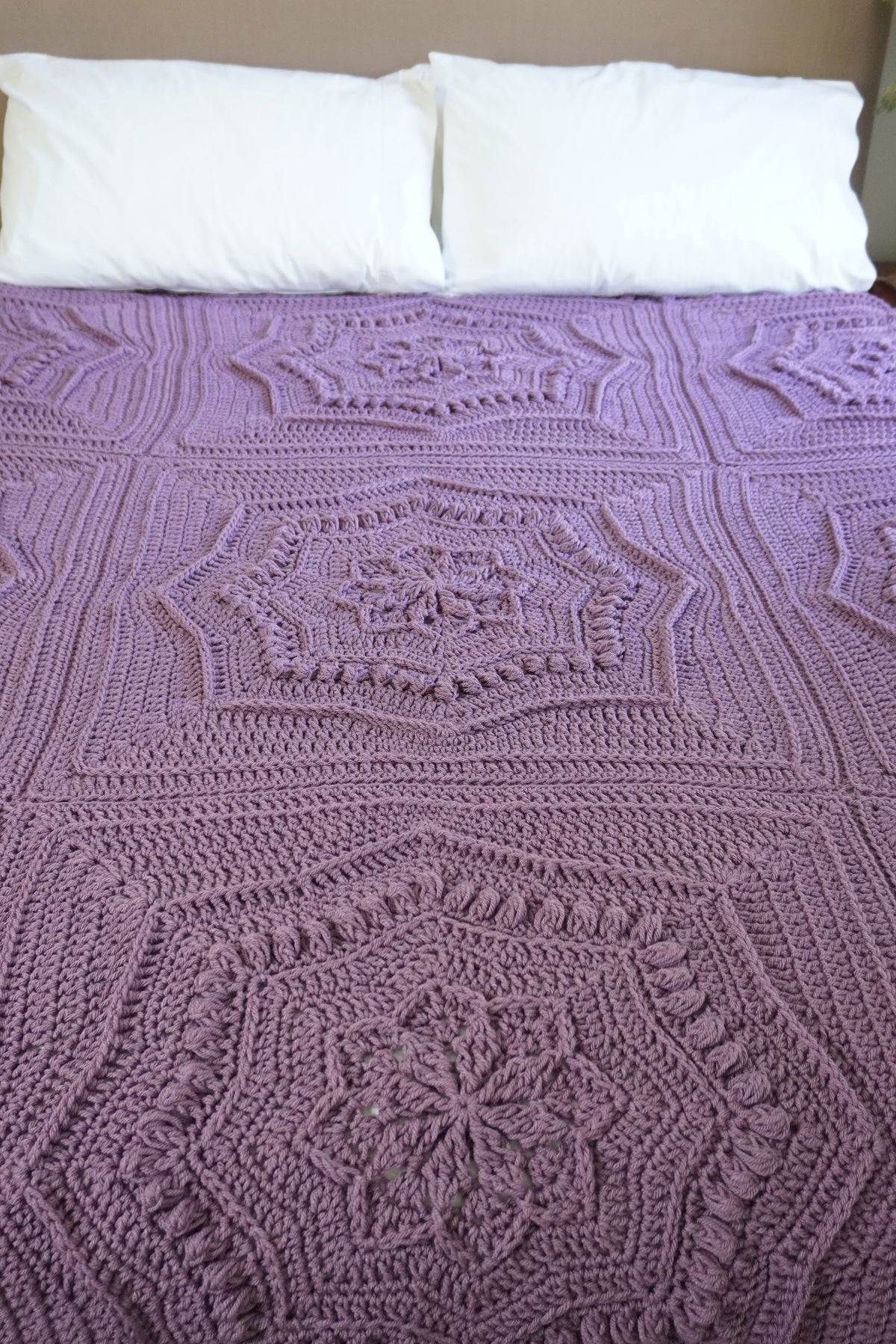A handmade crocheted blanket with a textured octagonal pattern in dusty purple, styled on a bed with two white pillows against a beige wall.