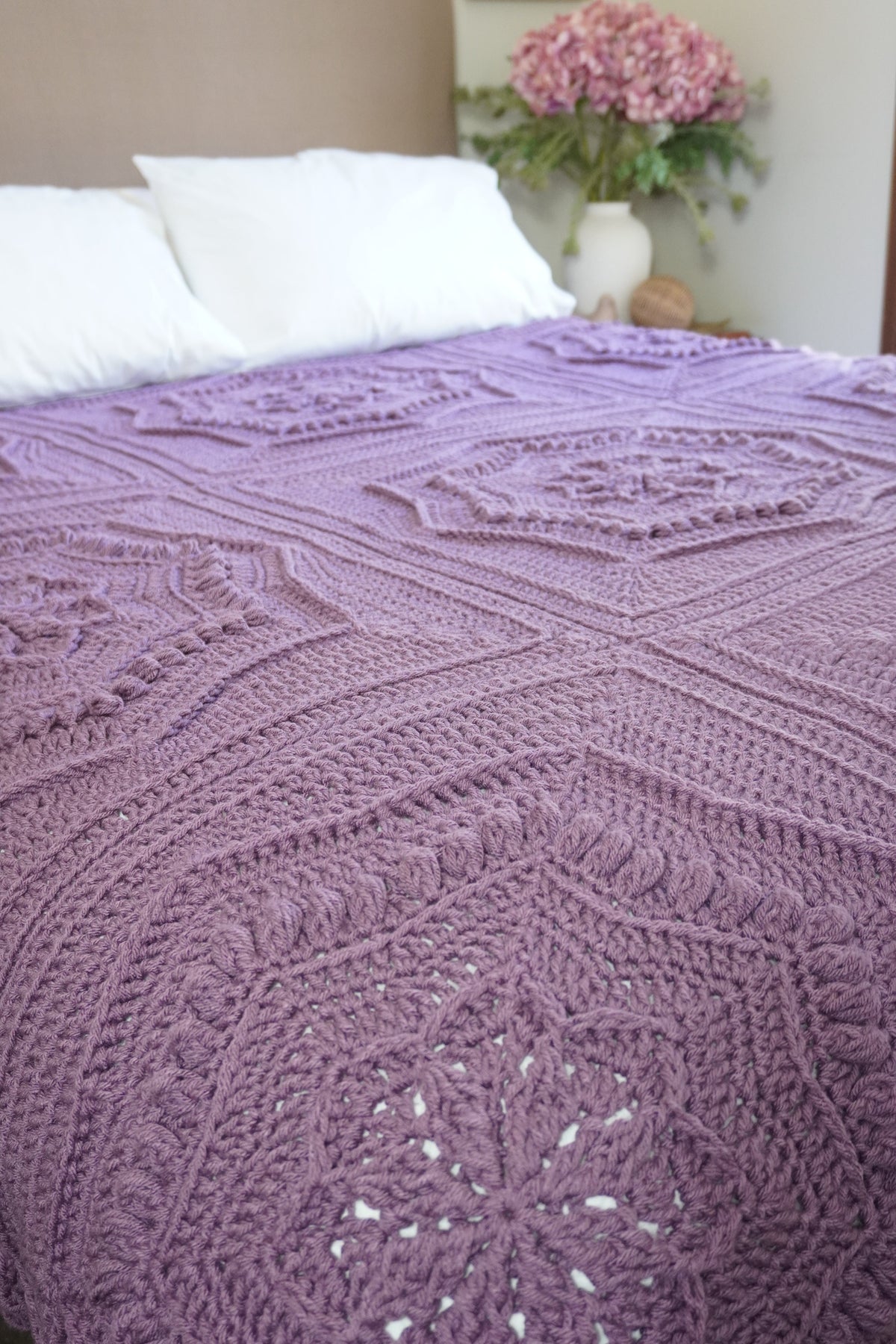 A lavender purple crocheted blanket with intricate granny square and floral lace patterns draped over a bed with white pillows in a bedroom setting.