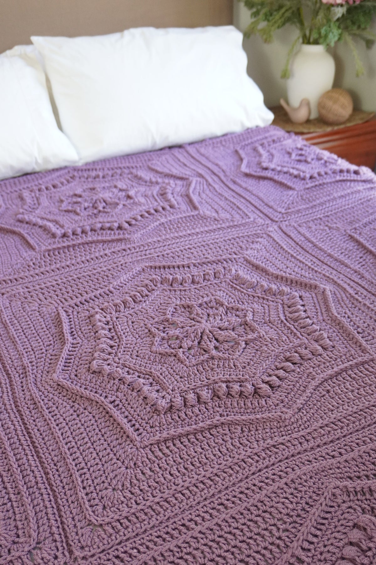 A handmade crocheted blanket in a dusty lavender-purple color, styled on a bed with white pillows and a bedside table in the background.