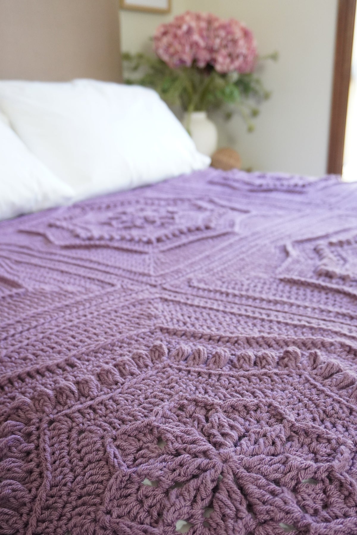 A close-up of a textured, crocheted blanket in a dusty purple/lavender color, spread across a bed with white pillows and a vase of pink flowers in the background.