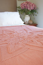 A peach-pink crocheted blanket with an intricate floral mandala pattern draped over a bed with white pillows, with a vase of pink flowers in the background.