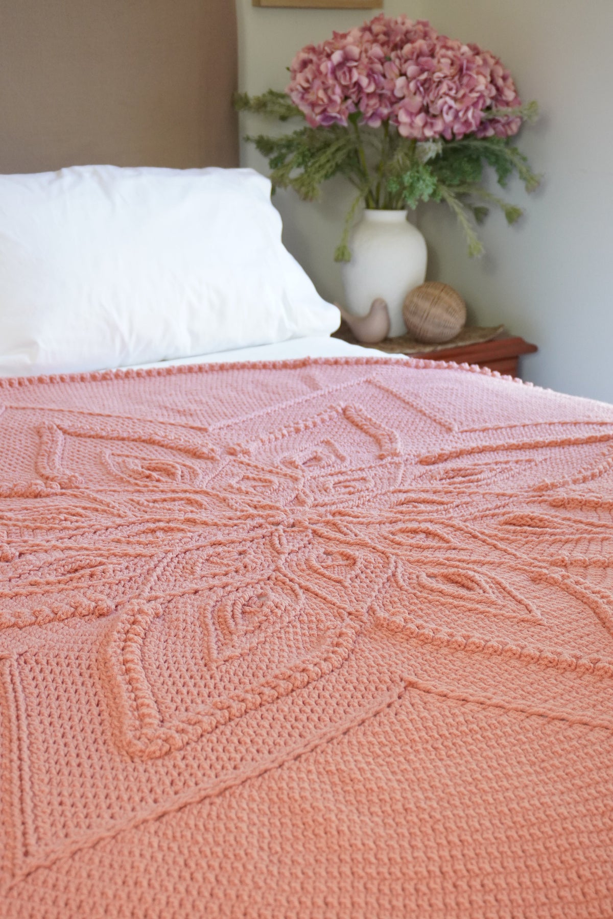 A peach-pink crocheted blanket with an intricate floral mandala pattern draped over a bed with white pillows, with a vase of pink flowers in the background.