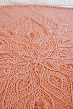 A close-up of a peach-pink crocheted blanket with an intricate floral and geometric pattern, laid out on a bed with white pillows visible at the top.