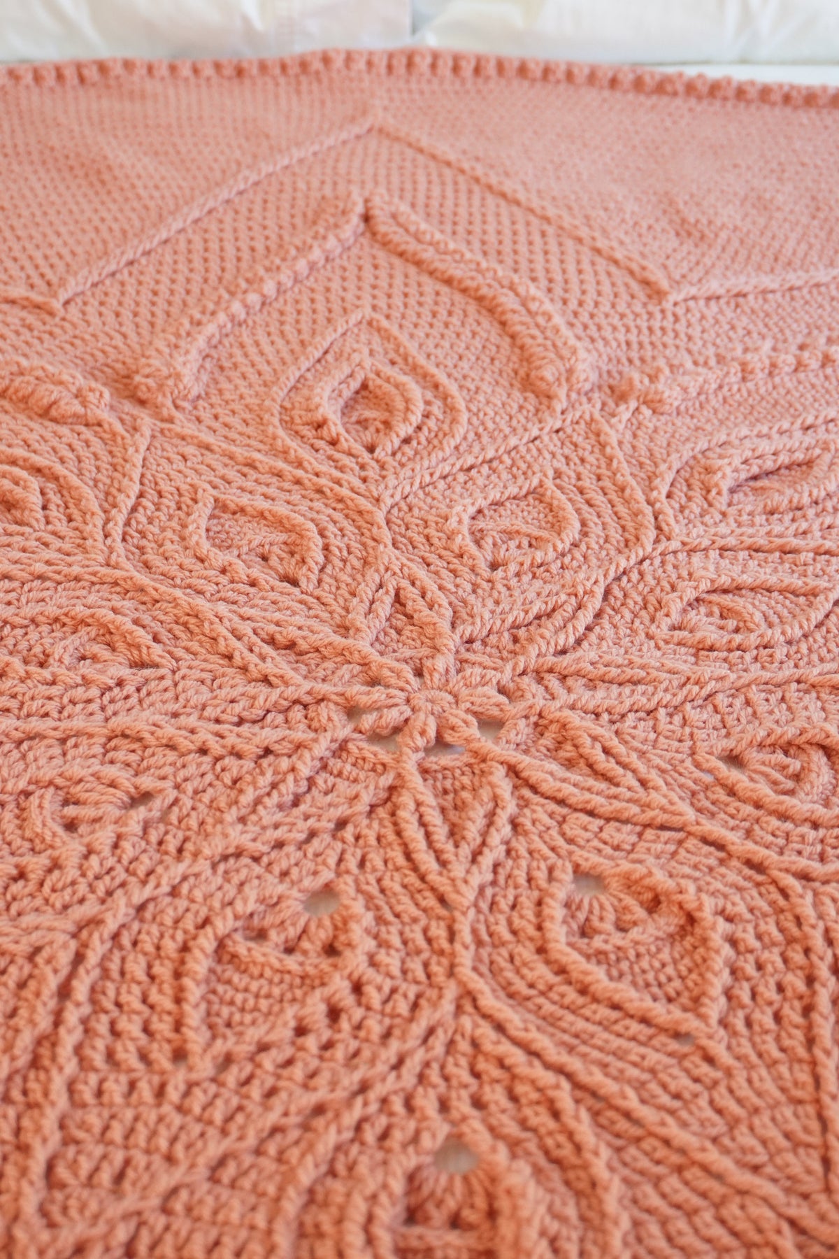 A close-up of a peach-pink crocheted blanket with an intricate floral and geometric pattern, laid out on a bed with white pillows visible at the top.