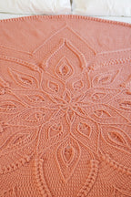 A peach-pink crocheted blanket with an intricate mandala pattern, spread out on a bed with white pillows visible at the top.